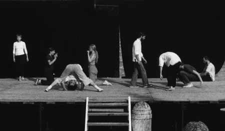 Scena plenerowa. Z tyłu sceny ściana z czarnej płachty. Po lewej stronie stoi aktorka ubrana w czarne spodnie i białą koszulkę z długim rękawem. Obok niej naprzeciwko siebie klęczą dwie aktorki. Pokazują sobie wnętrza dłoni. Pomiędzy nimi aktor lub aktorka wygląda, jakby robił/a fikołek. Z prawej strony czterech aktorów. Jeden z nich stoi, drugi jest pochylony, trzeci siada, czwarty siedzi z wyprostowanymi nogami.