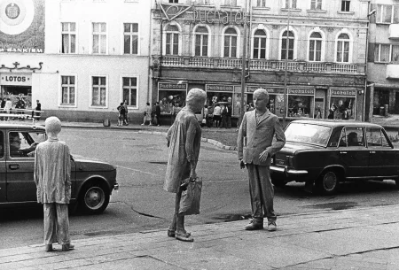 Przestrzeń miejska. Po ulica jadą samochody osobowe. W tle budynek z napisami "Puchatek". Na pierwszym planie, na chodniku stoją trzy postacie. Postać z lewej strony to dziecko odwrócone do widza plecami. Dwie postacie w centrum to kobieta i mężczyzna w trakcie rozmowy. Postacie wyglądają jak żywe rzeźby.