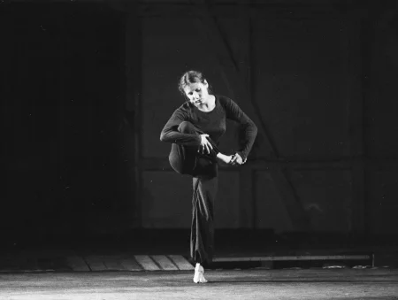 Aktorka stoi na pustej scenie w industrialnej przestrzeni. Ubrana jest w czarne spodnie i czarnę koszulkę z długim rękawem. Stoi na jednej nodze. Drugą nogę ma zgiętą, trzyma ją rękami w górze. Patrzy w dół.