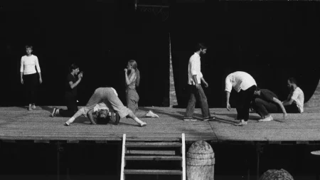 Scena plenerowa. Z tyłu sceny ściana z czarnej płachty. Po lewej stronie stoi aktorka ubrana w czarne spodnie i białą koszulkę z długim rękawem. Obok niej naprzeciwko siebie klęczą dwie aktorki. Pokazują sobie wnętrza dłoni. Pomiędzy nimi aktor lub aktorka wygląda, jakby robił/a fikołek. Z prawej strony czterech aktorów. Jeden z nich stoi, drugi jest pochylony, trzeci siada, czwarty siedzi z wyprostowanymi nogami.