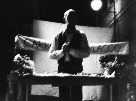 Niewyraźne zdjęcie. Aktor stoi zwrócony twarzą w stronę widza. Przed nim stoi kwietnik. Za jego plecami wisi biała szarfa z napisami.