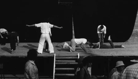 Scena plenerowa. Na scenę prowadzą drewniane schody. Z tyłu sceny ściana z czarnej płachty. Pod sceną przechodzi grupa widzów. Na scenie, po lewej stronie słabo widoczni aktor i aktorka. Stoją nisko pochyleni ku scenie. Obok nich stoi zwrócony plecami do widza aktor ubrany na biało. Aktor ma ręce wyciągnięte do boków. Po prawej stronie dwóch aktorów zwróconych do siebie twarzami kładzie się na scenę brzuchem do dołu. Za nimi stoi lekko pochylony aktor z głową zwieszoną w dół. Ubrany jest w jeansy i białą koszulę.