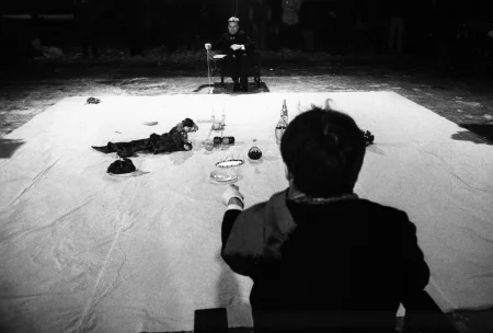 Na białej płachie leżą przedmioty, takie jak owoce, butelki i talerzyki. Po dwóch przeciwnych stronach płachty siedzi dwóch aktorów - jeden zwrócony plecami, drugi twarzą w stronę widza. Obaj trzymają w rękach napiętą linkę.