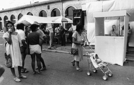 Ptrzestrzeń miejska. W małej białej kabinie stoi aktorka. Jest ubrana na biało i ma mikrofon przy twarzy. Obok kabiny przechodzi kobieta z wózkiem. Na ulicy stoi grupa ludzi.