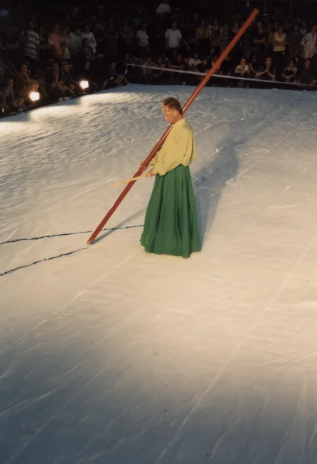 Scena ustawiona w przestrzeni miejskiej. Scena jest biała. Na scenie stoi aktor ubrany w zieloną spódnicę i jasnozieloną koszulę. W ręce trzyma długi czerwony kij, z którego wysypuje się proszek. W tle widać publiczność.