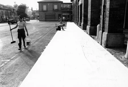 Przestrzeń miejska. Grupa aktorów i aktorek rozwija na ulicy między budynkami wielką biała płachtę. Po lewej stronie przechodzi aktorka z wiadrem i szczotką.