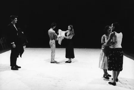 Dwóch aktorów i trzy aktorki na białej płachcie. Aktor po lewej stronie stoi zwrócony profilem w stronę widza. Ubrany jest w czarny garnitur. W centrum aktor i aktorka wyglądają, jakby wymieniali się ubraniami. Z prawej strony stoją aktorki, które rozmawiają i uśmiechają się.