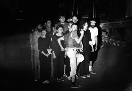 Bardzo blisko siebie stoją aktorzy i aktorki. Są statyczni. Przed nimi przechodzi zwrócony profilem do widza aktor. Wysoko unosi nogi i macha rękami. Patrzy w bok.