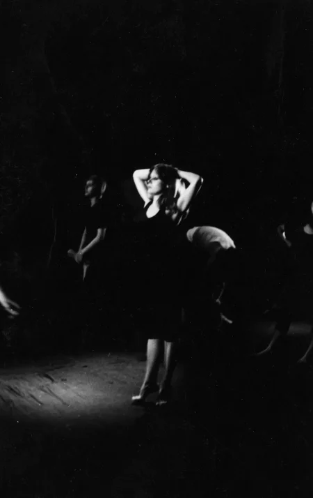 Rozmazane zdjęcie. W centrum stoi aktorka ubrana na czarno. Ma ręce założone na tył głowy. Za nią stoi kilku słabo widocznych aktorów.