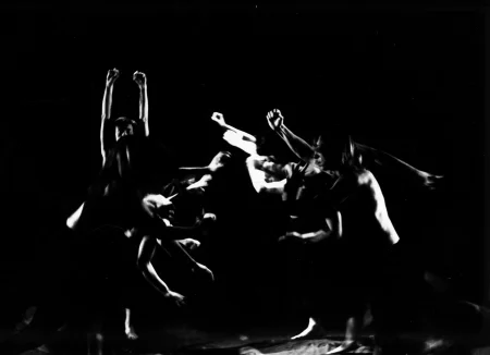 Po lewej stronie stoi aktor. Jest wyprostowany i ma wyciągnięte do góry ręce. W centrum grupa aktorów w ruchu. Idą przed siebie i trzymają w górze zaciśniętą pięść.