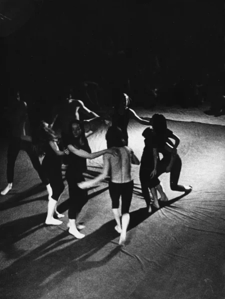 Scena wyglądająca jak wyłożona prześcieradłem. Na scenie grupa aktorów i aktorek widzianych od góry.Stoją w okręgu i wyglądają, jakby tańczyli. Aktorzy ubrani są w ciemne spodenki, mają nagie torsy. Aktorki ubrane są w czarne spodenki i czarne podkoszulki. Na drugim planie siedząca grupa widzów.