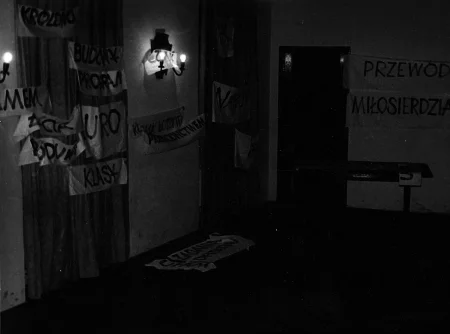 Pomieszczenie w budynku. Ławka i stół. Na tylnej ścianie drewniane drzwi. W pomieszczeniu na białym materiale wiszą wymalowane czarną farbą hasła. Najlepiej widoczny jest napis "PRZEWÓD MIŁOSIERDZIA".