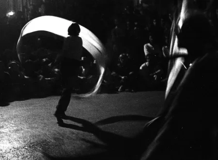 Niewyraźne zdjęcie. Aktorka zwrócona plecami do widza macha białą wstęgą. Przed nią siedzą widzowie.