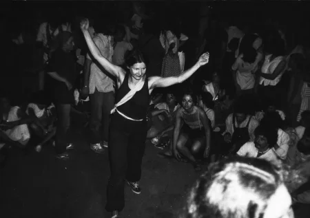 Grupa widzów i aktorów. Trudno rozsztrzygnąć, kto jest kim. W centrum idzie uśmiechnięta kobieta z wyciągniętymi w górę rękami. Ubrana jest w czarne trampki, czarne spodnie i czarną koszulkę na ramiączka.