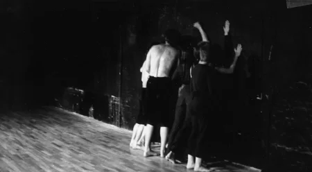 Grupa aktorów i aktorek zwróconych plecami w stronę widza stoi pod ścianą. Aktorzy i aktorki uderzają w ścianę.