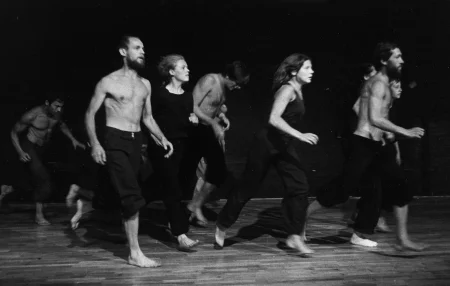 Grupa aktorów i aktorek biegnie po panelowej podłodze. Aktorzy ubrani są w czarne spodenki, mają nagie torsy. Aktorki ubrane są w czarne spodenki i czarne podkoszulki. Wszyscy są boso.