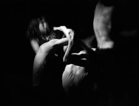 Niewyraźne, rozmazane zdjęcie. Dwóch aktorów i jedna aktorka trzymają się za ręce. Prawdopodobnie klęczą na podłodze. Aktorka ma na sobie koszulkę, aktorzy mają nagie torsy. Po prawej stronie od nich widać nagi, wyprostowany tors aktora.