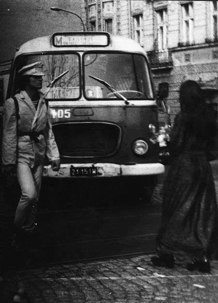 Centralnie stoi bus ustawiony maską w stronę widza. Z lewej strony idzie aktor. Ubrany jest w jasne spodnie, jasny krótki płaszcz i jasną czapke. Z prawej strony, odwrócona plecami do widza idzie aktorka. Ubrana jest w czarną sukienkę. W tle widać budynek mieszkalny.