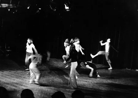 Rozmazane zdjęcie. Grupa aktorów i aktorek wygląda, jakby tańczyła. Ich ruchy są bardzo ekspresyjne. Wszyscy mają na sobie jeansy. Niektórzy mają na sobie koszulki, inni mają nagie torsy. Z sufitu zwistają reflektory. Na pierwszym planie kilkoro widzów siedzi na widowni i ogląda aktoró.