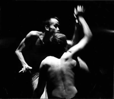 Dwoch aktorów stoi naprzeciwko siebie. Jeden z nich jest zwrócony do nas tyłem, drugi przodem. Obaj nie mają koszulek, są przygarbieni i wyciągają jedną rękę do góry. Sprawia to wrażenie, jakby przeglądali się w lustrze.