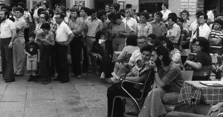 Pokaźna grupa ludzi, prawdopodobnie widzów i widzek teatralnych. Prawdopodobnie oglądają spektakl. Są w różnym wieku. Mają ubrania typowe dla mody lat siedemdziesiątych - jeansy i koszule lub koszulki wkasane w spodnie. Niektórzy stoją, inni siedzą przy stolikach.
