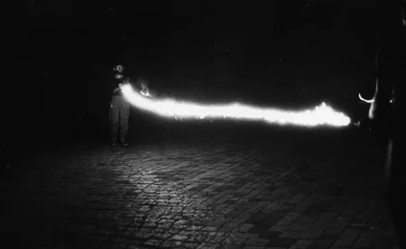 Niewyraźne zdjęcie. Przestrzeń miejska. Na brukowanej ulicy stoi dwóch aktorów. Trzymają w rękach przedmiot emitujący silne światło.