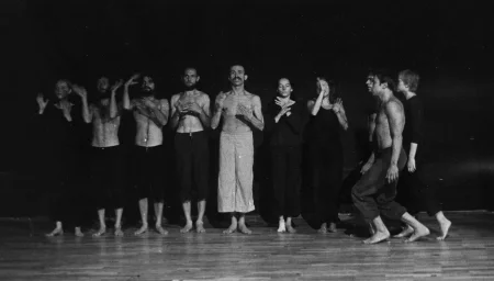 Grupa aktorów i aktorek stoi w rzędzie wzdłuż sceny twarzą do widza.. Gestykulują dłońmi.Pośrodku stoi aktor w jasnych spodniach, jego tors jest nagi. Aktor jest bosy. Ma wąsy i wygląda, jakby coś mówił. Aktorzy i aktorki po jego lewej i prawej stronie ubrani są w czarne spodnie. Aktorzy mają nagie torsy, aktorki czarne koszulki. Od prawej strony zwróceni profilem do widza idzie dwóch aktorów i jedna aktorka. Są lekko przygarbieni. Aktorzy są ubrani w czarne spodnie, mają nagie torsy. Aktorka ubrana jest w czarne spodnie i czarną koszulkę. Są boso. Podłoga, na której stoją aktorzy, wygląda na drewnianą.