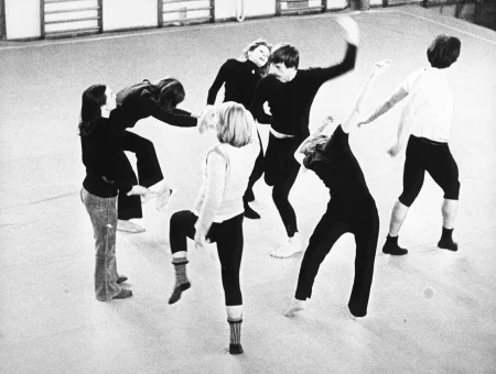 Siedmioro aktorów i aktorek na sali gimnastycznej. Wyglądają, jakby tańczyli. Wszyscy ubrani są w ciemne spodnie. Pięcioro z nich ubrana jest w czarne koszulki. Pozostała dwójka ubrana jest w białe koszulki.