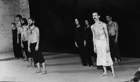 Grupa aktorów i aktorek stojących na tle czarnej płachty. Aktorzy ubrani są w czarne spodenki, mają nagie torsy. Aktorki ubrane są w czarne spodnie i czarne koszulki. Wśród grupy wyróżnia się aktor ubrany w jasne spodenki. Wszyscy są boso. Wszyscy delikatnie przechylają się w tę samą stronę.