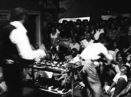 Niewyraźne zdjęcie. Aktor zwrócony plecami do widza przemawia do roześmianej publiczności. Obok niego stoi kwietnik. Wsród widzów stoi aktor, który wygląda, jakby dawał im coś do powąchania.