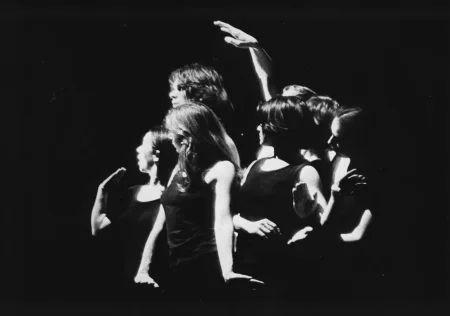 Grupa aktorek i aktorów zwróconych do nas przodem patrzy w prawo. Wykonują gesty rękami. Nad głową jednej z aktorek widoczna jest wyciągnięta ku górze, prawdopodobnie męska ręka.