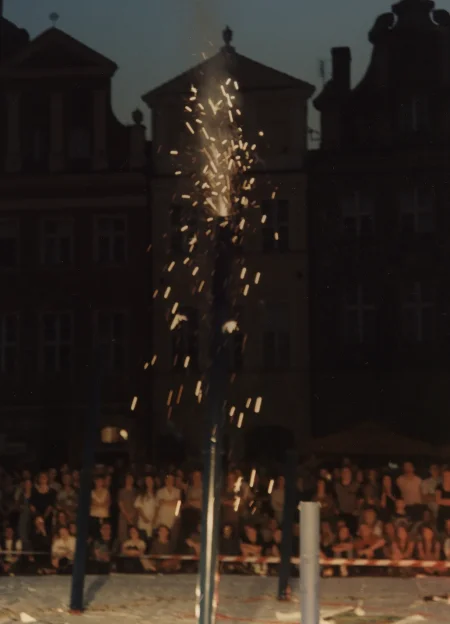Scena ustawiona w przestrzeni miejskiej. Na scenie stoi duży fajerwerk, z którego wyskakują iskry. W tle widać budynki i publiczność.