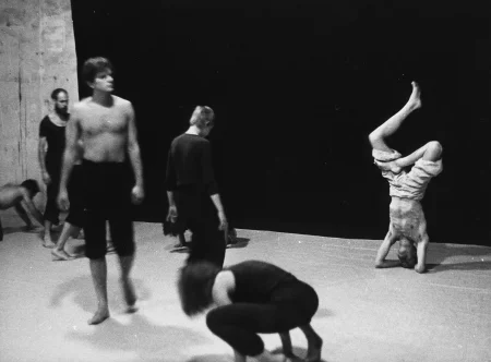 Grupa aktorów i aktorek wykonuje różne ruchy gimnastyczne. Nie zwracają na siebie nawzajem uwagi. Aktor po lewej stronie stoi i patrzy w przestrzeń. Ubrany jest w czarne spodenki, ma nagi tors. W centrum, na pierwszym planie pochylona aktorka podpiera się dłońmi o podłogę. Ubrana jest w czarne spodnie i czarną koszulkę na ramiączka. Za nią stoi zwrócona tyłem do widza aktorka. Ubrana jest w czarne spodnie i czarną koszulkę. Po prawej stronie, w głębi aktor stoi na głowie. Ubrany jest w jasne spodenki, ma nagi tors. Ma twarz zwróconą w stronę widza. Na tylnej ścianie zwisa czarna płachta będąca tłem dla działań aktorów.