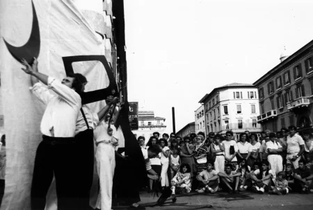 Przestrzeń miejska. Po lewej stronie pod białą płachtą grupa aktorów i aktorek trzyma w rękach trudne do zidentyfikowania czarne dwuwymiarowe rekwiyty. Rekwizyty wyglądają na wycięte z papieru. Po prawej stronie stoi duża grupa widzów oglądających aktorów. W tle widać budynki.