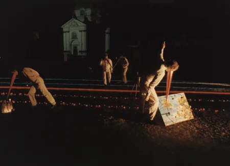 Niewyraźne zdjęcie. Noc. Przestrzeń miejska. Noc. Przestrzeń miejska. Na piasku leżą trasy wyznaczone przez czerwone i niebieskie lampki. Pośród lampek stoją aktorzy i aktorki ubrani na biało. Do twarzy mają doczepione długie czerwone dzioby. Obok jednego z aktorów leży obraz przedstawiający niebo i drzewa. W tle widać biały budynek.