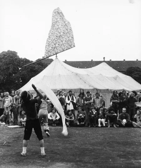 Plener. Aktor podrzuca wysoko flagę w kwiaty. W tle siedzą i stoją widzowie. Na dalszym planie stoi namiot cyrkowy.