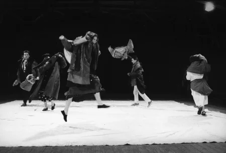 Grupa aktorów i aktorek na białej płachcie. Mają na sobie wiele warstw ubrań wyglądających na założone przypadkowo. Aktorzy i aktorki skaczą i biegają.