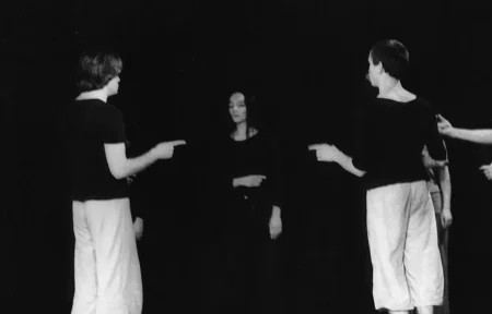 Grupa aktorów i aktorek stoi w okręgu. Wskazują na siebie palcami. Dwóch aktorów na pierwszym planie jest odwróconych plecami w stronę widza. Ubrani są w jasne spodnie i czarne koszulki. Na drugim planie stoi aktorka ubrana na czarno.