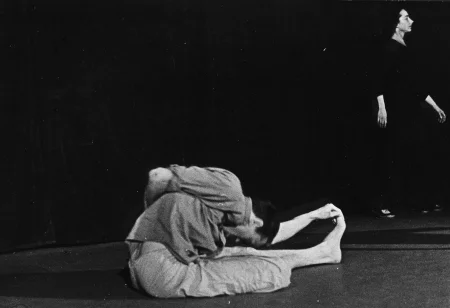 Aktor, zwrócony profilem do widza, siedzi na podłodze. Ubrany jest w jasne spodnie i jasną koszulę. Jest boso. Ma wyprostowaną lewą nogę. Lewą dłonią trzyma palce stopy. Głową sięga kolan. Po prawej stronie stoi aktorka. Jest ubrana na czarno.