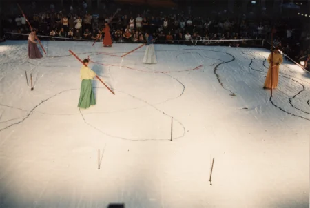 Duża biała scena ustawiona w przestrzeni miejskiej. Scena wygląda, jakby był na niej śnieg. Na scenie stoi czterosoobowa grupa aktorów i aktorek ubranych w kolorowe suknie w stylu azjatyckim. Trzymają w rękach długie czerwone kije i rysują wzory na scenie. Pod sceną stoi grupa widzów.