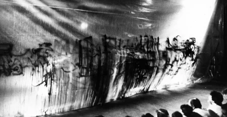 Płachta z nieczytelnymi napisami namalowanymi farbą. Płachtę ogląda ledwo widoczna publiczność zwrócona do widza plecami.