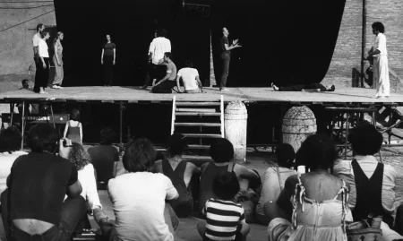 Scena plenerowa. Na pierwszym planie odwróceni plecami widzowie patrzą na scenę. Na scenę prowadzą drewniane schody. Z tyłu sceny ściana z czarnej płachty. Pośrodku, zza płachty wystaje latarnia. Na scenie stoją aktorzy i aktorki w różnych działaniach. Wyglądają, jakby mieli na sobie prywatne ubrania. Wspólnie tworzą choreograficzną kompozycję. Na dalszym planie, za sceną widać budynki.