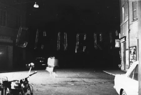 Niewyraźne zdjęcie. Przestrzeń miejska. Na ulicy między budynkami stoją motory i samochody. Nad ulicą wisi trudny do rozczytania napis.