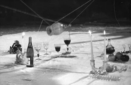 Na białej łachcie leżą przedmioty, takie jak butelki, kieliszki, świece, kwiaty i owoce. Przedmioty są obwiązane sznurkami. Nad nimi na sznurkach unosi się butelka, z której do jednego z kieliszków leje się wino.