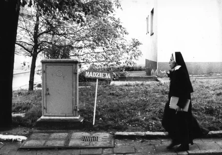 Przestrzeń miejska. Na trawniku, obok studzienki kanalizacyjnej, stoi strzałka z napisem "NADZIEJA". Obok strzałki, chodnikiem przechodzi zakonnica.
