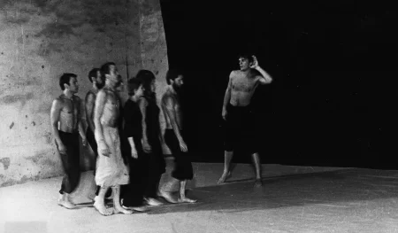 Na tle czarnej płachty przechodzi zwrócona profilem do widza grupa aktorów i aktorek. Aktorzy ubrani są w czarne spodnie i mają nagie torsy. Aktorki ubrane są w czarne spodnie i czarne koszulki. Wyróżnia się jeden aktor, który ubrany jest w jasne spodenki. Wszyscy są boso. Obok grupy stoi aktor. Trzyma dłoń przy uchu, jakby czegoś nasłuchiwał.