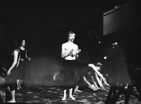 W centrum sceny stoi aktor. Ubrany jest w czarne spodenki, jego tors jest nagi. Aktor jest boso. Ma obie ręce zgięte i wyciągnięte przed siebie. Wokół niego aktorki wykonują ekpresyjne ruchy. Ubrany są w czarne spodnie i czarne koszulki. Są boso. Scena, na której stoją aktorzy, jest czarna ma białe plamy.