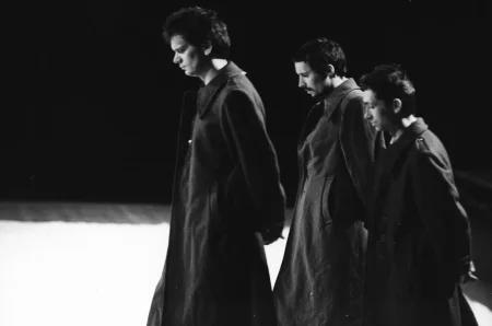 Trzech aktorów zwróconych profilem w stronę widza. Wszyscy ubrani są w długie ciemne płaszcze. Mają poważny wyraz twarzy.