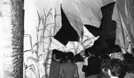 Przestrzeń miejska. Uliczką idzie tłum ludzi zwrócony plecami w stronę widza. Przechodzą przez płachtę, rozrywając ją. Na płachcie namalowane są rośliny przypominające trzciny.