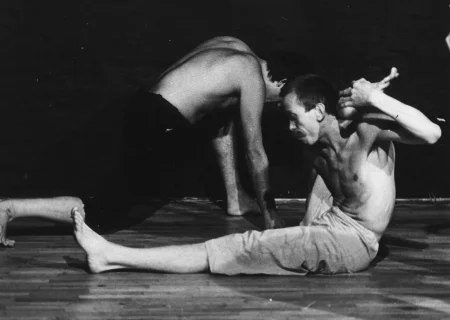 Aktor, zwrócony profilem do widza, siedzi na podłodze. Ubrany jest w jasne spodenki, ma nagi tors. Jest boso. Jedną nogę ma wyprostowanę, drugą trzyma sa swoją głową. Za aktorem klęczy podparty rękami o podłogę drugi aktor. Ubrany jest w czarne spodenki, ma nagi tors. Jest słabo widoczny.